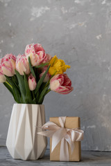 Colorful pink and yellow tulips in a white vase on the table on a light gray background, box with a gift. Kraft packaging and a beautiful bow.
