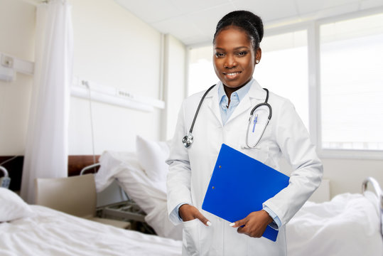 medicine, profession and healthcare concept - smiling african american female doctor in white coat with clipboard and stethoscope over hospital ward background