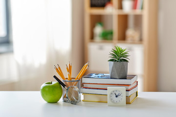 education and school concept - books, magnifier, pencils, plant in flower pot with green apple and alarm clock on table at home