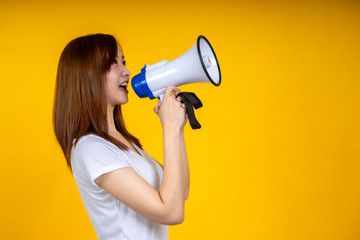 Young Asian woman in white casual t-shirt looking aside, scream in megaphone isolated on bright yellow wall background in studio. People lifestyle concept. Mock up copy space