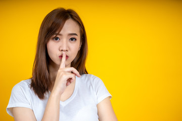 Young Asain woman requires silence. Young beautiful brunette has put forefinger to lips as sign of silence, isolated on yellow
