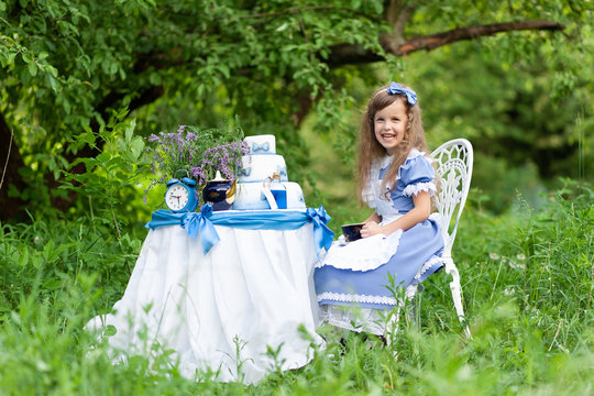 A little cute girl in the costume "Alice from Wonderland" holds a tea party at her magic table. Photographed in nature.