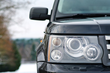 Modern luxury black premium SUV right car headlight with washer closeup over winter forest with snow background with copy space. Concept of expensive luxury premium sports offroad SUV.
