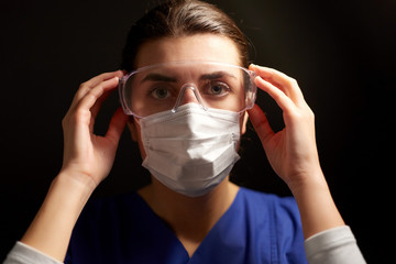 health, medicine and pandemic concept - young female doctor or nurse wearing goggles and face protective mask for protection from virus disease over black background