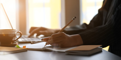 Cropped image of woman's hands writing on notebook and typing on laptop while sitting at the black...