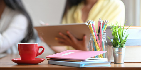 Red ceramic coffee cup putting and small dish putting on wooden desk that surrounded by notebook, stack of books. potted plant, pencils in glass vase over graphic designer team as background.