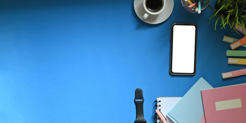 Top view image of white blank screen smartphone putting on colorful working desk that surrounded by marker pens, notebooks, smartwatch, coffee cup, pencil holder and potted plant. Cluttered workspace.