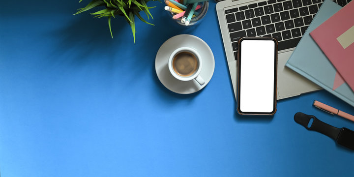Top view image of white blank screen smartphone putting on colorful working desk that surrounded by marker pens, notebooks, smartwatch, coffee cup, pencil holder and potted plant. Cluttered workspace.