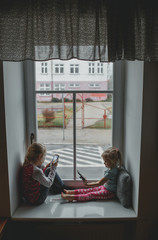 two sisters play a mobile phone on the sill of a large window. girls play with gadgets and have...