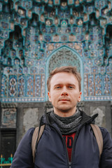 A young blond man in a jacket and with a backpack stands against the background of a Muslim mosque in St. Petersburg, a beautiful tile, mural, pattern