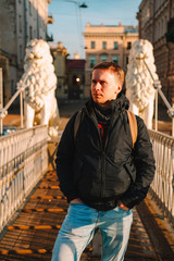 A young blond man in a jacket and with a backpack stands on the bridge with statues of white lions...