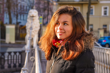 A brunette girl with long hair in a jacket stands on a bridge with statues of white lions in the middle of the canals of St. Petersburg, Attractions, morning spring