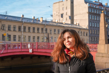 Fototapeta na wymiar A girl with long hair in the middle of the street against the background of the Sights of St. Petersburg, a beautiful historical building near the red bridge near the river channel, morning spring