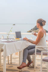 A woman sitting at a beach restaurant having breakfast while working on a laptop.