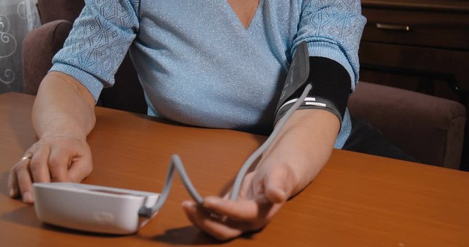 Close up detail shot of hands. Elderly European woman in blue sweater measuring her blood pressure by herself on brown wooden table using white electronic automatic blood pressure monitor at home. 