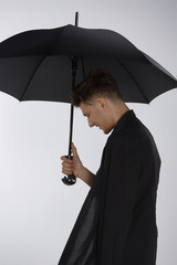 A young dark-haired man in a black raincoat is holding a large black umbrella with an unique silver...