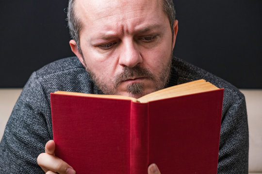 Close-up of serious young bearded man reading red book on black background
 - Powered by Adobe