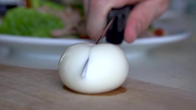 Male hand cuts semi hard boiled egg with kitchen knife on wooden chopping board revealing beautifully yellow egg yolk inside, SLOW MOTION.