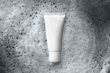 Top down view closeup organic gentle facial skincare product cleanser white tube with a blank label on white bath foam bubbles texture as a background.