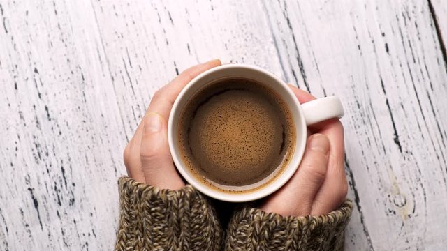 Woman Wearing Sweater Holding A Cup Of Coffee. Stirred Drink With Brown And Orange Foam And Bubbles Rotating In The Cup. Top Down Slow Motion Shot, 4K