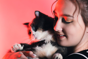 portrait of a young happy woman with a cat in her arms in red color lighting in the studio, girl plays with pet, lovely animals