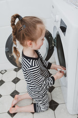 Cute girl in striped clothes puts things into the washing machine. Cleanliness and Household Concept