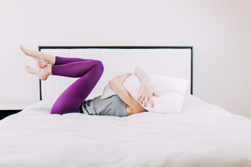 Young woman at home while quarantine, do sports while self-isolation, home yoga, woman in purple leggins on the bed, cry into pillow, woman hugging pillow