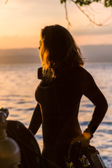 Female Scuba Diving Instructor Wearing a Wet Suit Standing Next to a Twin Tank at Sunset