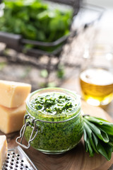 Wild leek pesto with olive oil and parmesan cheese in a glass jar on a wooden table. Useful properties of ramson. Leaves of fresh ramson. Vertical
