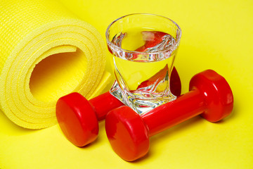 Fototapeta na wymiar red dumbbells, a glass of water, a yellow rug, colored background, sports, energy drink, equipment for the gym