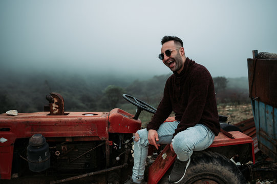Armenian Caucasian man sits on the old broken red tractor in the mountains during the foggy day. Finding the interesting abandoned things while travelling. Travel concept. Natural skin tone