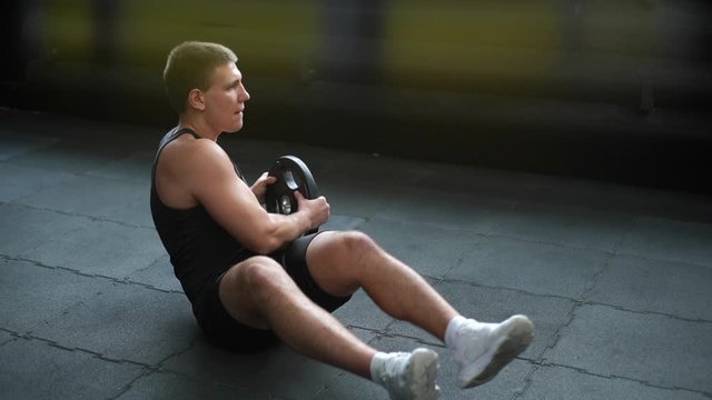 Muscular strong man doing russian twist exercise with weight from barbell on black mat during sport workout training in modern dark gym. Concept of healthy lifestyle. Shooting in slow motion.