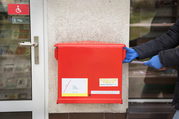 A man wearing a protective mask and gloves tosses a letter into a red mailbox. There is a pandemic coronavirus in 2020
