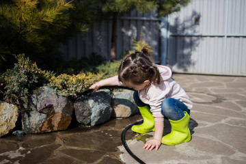 Fototapeta na wymiar A girl in green rubber boots is watering the garden. A girl in a pink jacket and jeans pours water from a hose on plants and firs