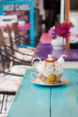 Tea pot set for one on bright turquoise table at an outdoor tea-room. Background out of focus bright pink, purple and blue.