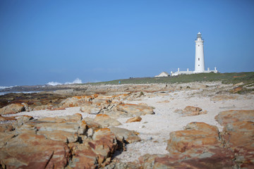 A white lighthouse in the distance with grass sand and rocks in the foreground. To the left of the lighthouse a wave crashes against rocks with an explosive spray.