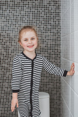 Cute girl in striped clothes is standing on the toilet. Black and white interior in fine tiles. TThe idea of a child’s activity during quarantine