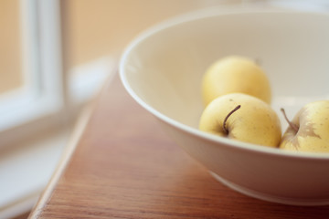 Golden delicious apples in a white ceramic bowl on a wooden table next to a window. Soft out of focus background of white and cream tones.