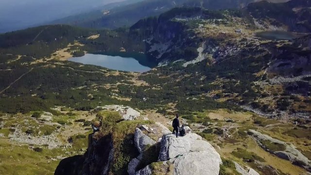 Aerial, tilt down, drone shot over hikers sitting on a rocky mountain ledge, sunny day, in Rila National Park, Bulgaria
