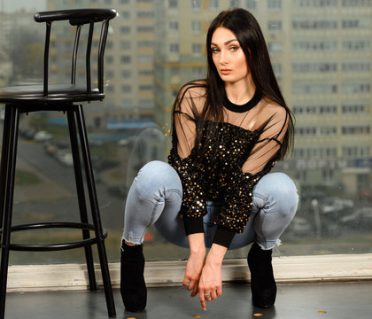 Photo model glamorous girl in brunette jeans posing on a chair against the background of a window