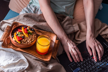 Healthy breakfast in bed. A young woman in pajamas is typing on laptop. A tray with a bowl of granola, fruits, glass with orange juice. Modern sunny morning. Self isolation. Top view.