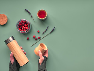 Zero waste tea in travel flask. Making herbal infusion in eco friendly insulated bamboo flask with fresh cranberry tea. Trendy flat lay with hands holding the flask and bamboo cups, copy-space.