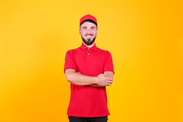 Young handsome delivery guy wearing red uniform and cap holding posing over isolated yellow background. Portrait of friendly bearded man in polo shirt and baseball hat. Copy space for text.