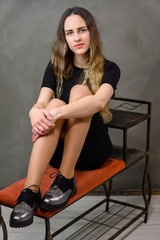 Fototapeta premium Vertical studio portrait of a caucasian girl in a black dress with long hair sitting on a stand on a gray background.