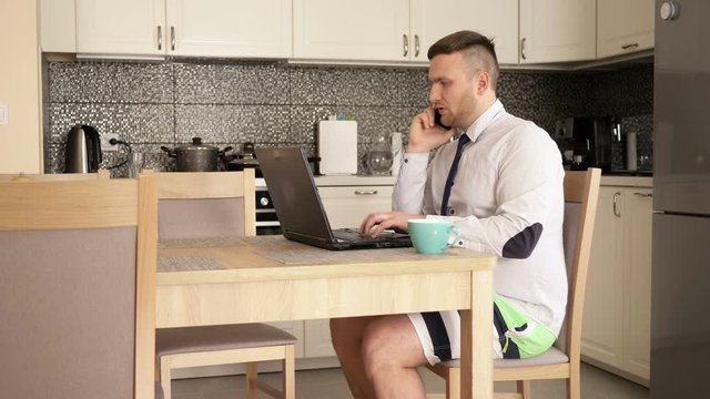 Home Office. Young businessman is working intensively while sitting at the kitchen table. He communicates simultaneously on skype and mobile phone. For comfort, dressed in shorts.