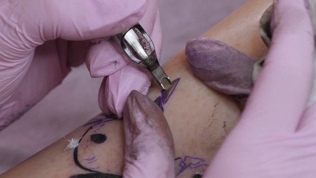 Master doing tattooing in tattoo studio. Professional tattooist works in studio. A woman in pink gloves makes a tattoo on the leg of a young girl. Slow motion.