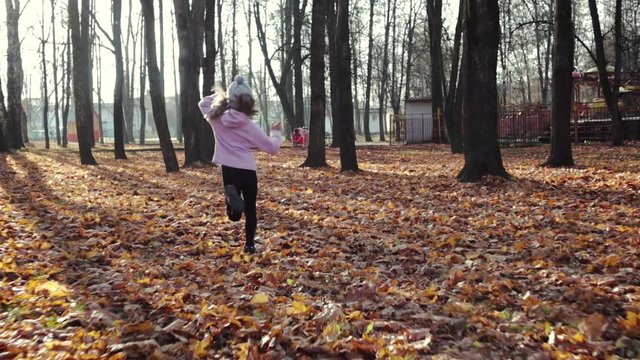 Steadicam shot a rear view of a young pretty schoolgirl in black leggings and a pink hoodie running through dry fallen leaves in an autumn Park. Happy girl playing in the fresh air