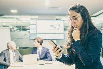 Thoughtful businesswoman sitting on table and looking at phone. Focused young woman in eyeglasses using smartphone. Business, technology concept