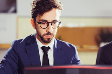 Fototapeta premium Focused young man in eyeglasses looking at laptop. Front view of concentrated young manager working with laptop in office. Business, working late concept