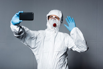 Fototapeta na wymiar man in personal protective equipment taking selfie and waving hand on grey